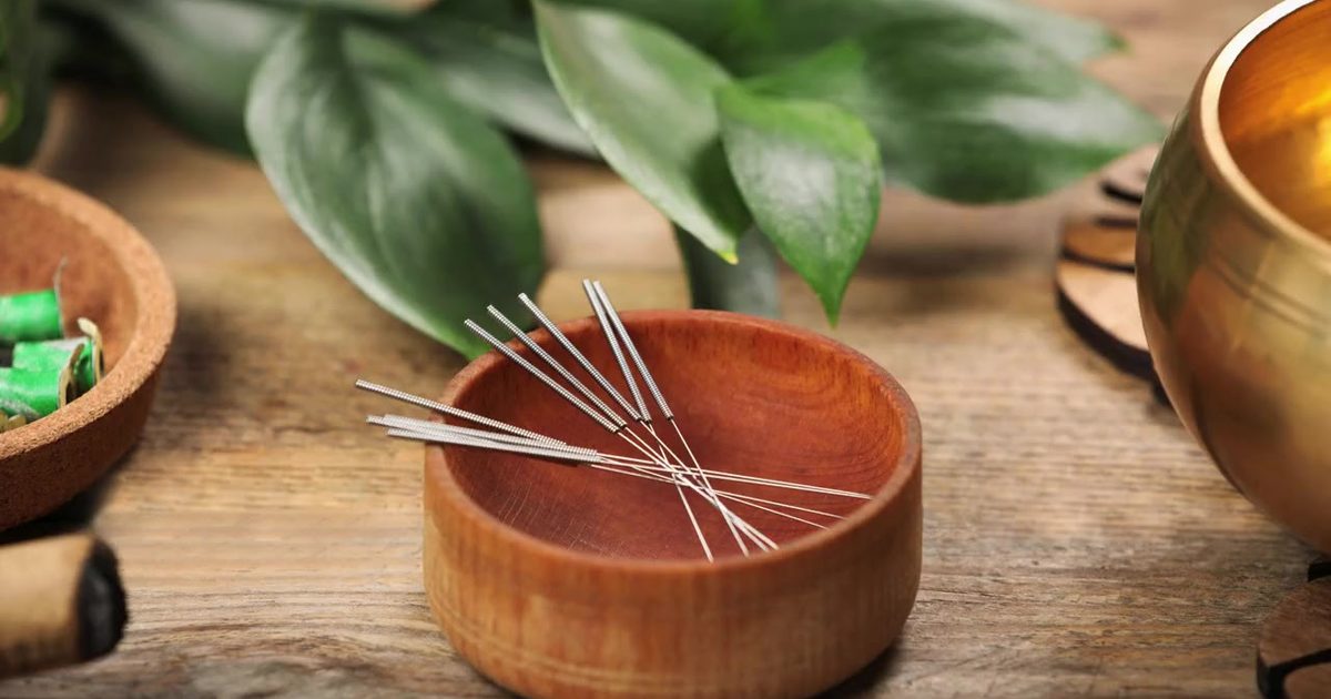 Fine acupuncture needles resting in a small wooden bowl beside green leaves