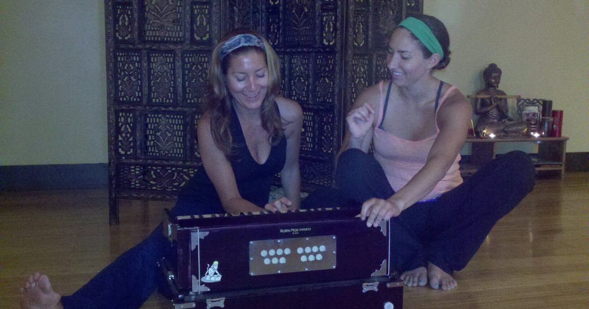 Two women singing together at a harmonium during a kirtan gathering