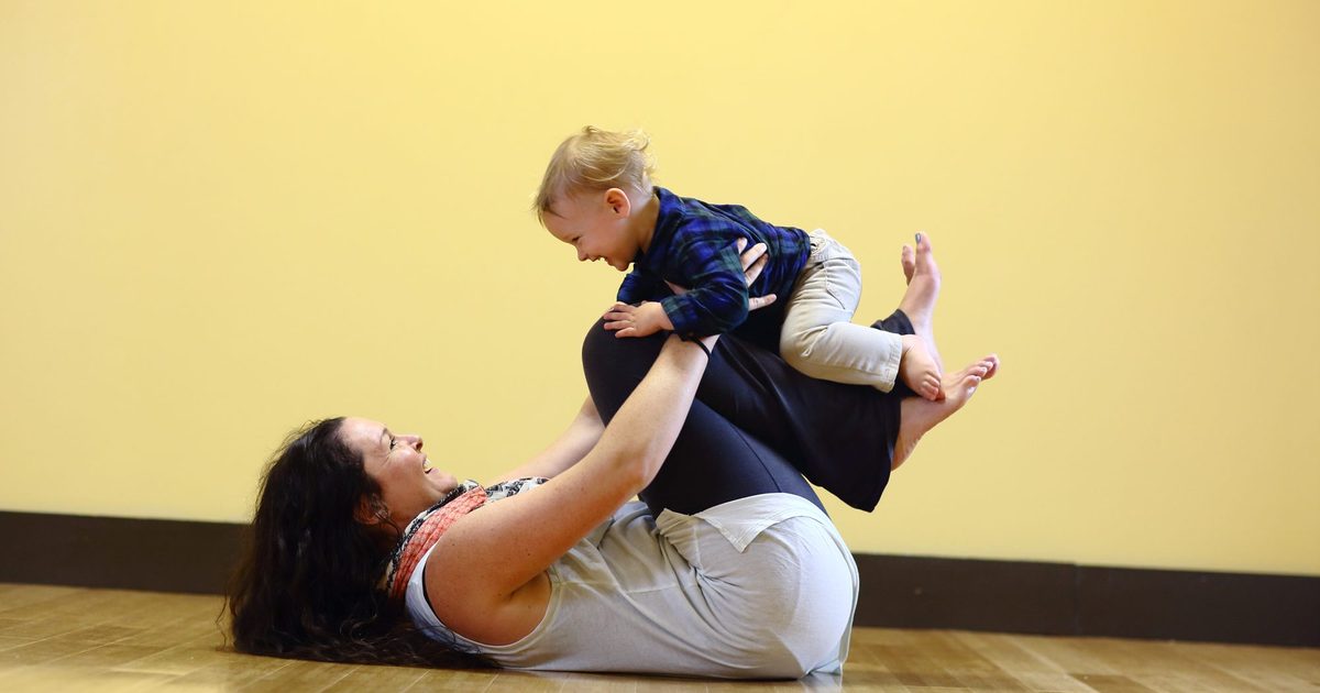 A mother lying on her back lifting her laughing baby on her shins in a postnatal yoga class